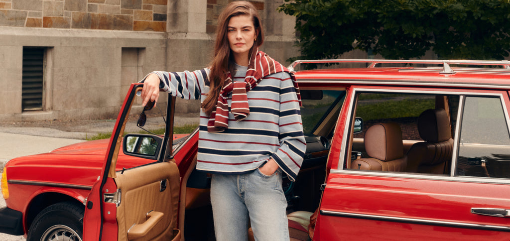 Woman opening a car door in a blue striped tee and jeans with a red striped tee draped over her shoulders. 