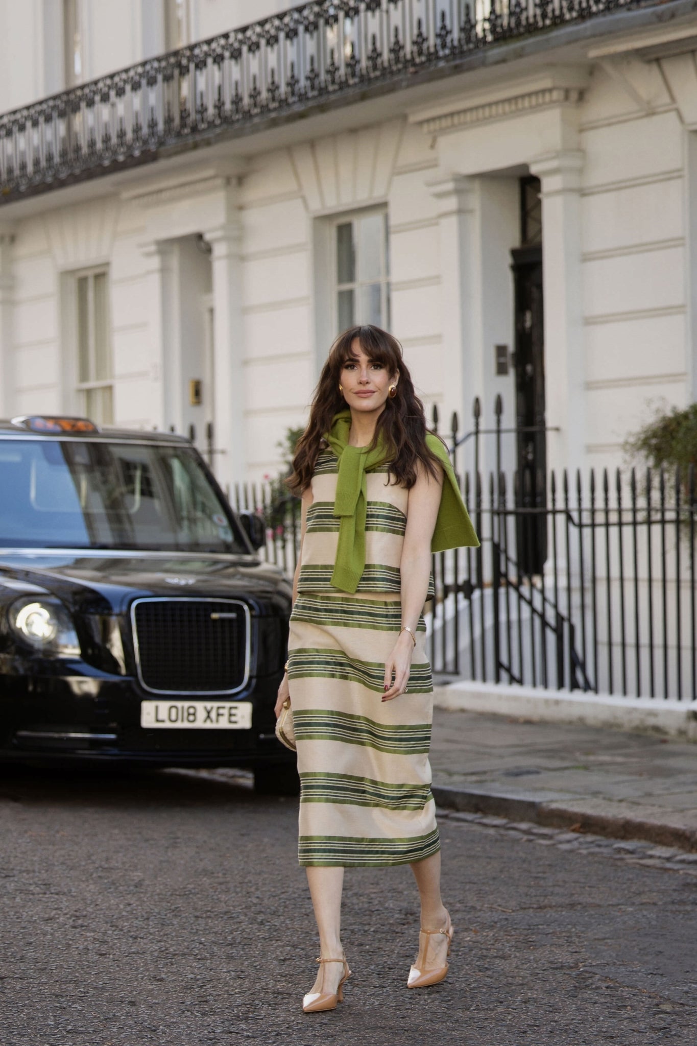 Woman in classic beige and green-striped Satin Stripe Ellington Skirt, effortlessly styled with a draped sweater, embodying high-end, fun elegance on a cobblestone street.