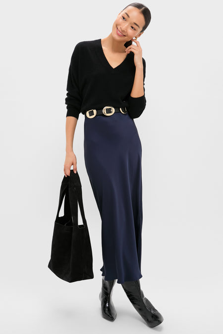 A woman smiles in a Tuckernuck oversized black cashmere V-neck sweater, navy blue satin skirt with wide belt, black boots, and carries a black tote bag.