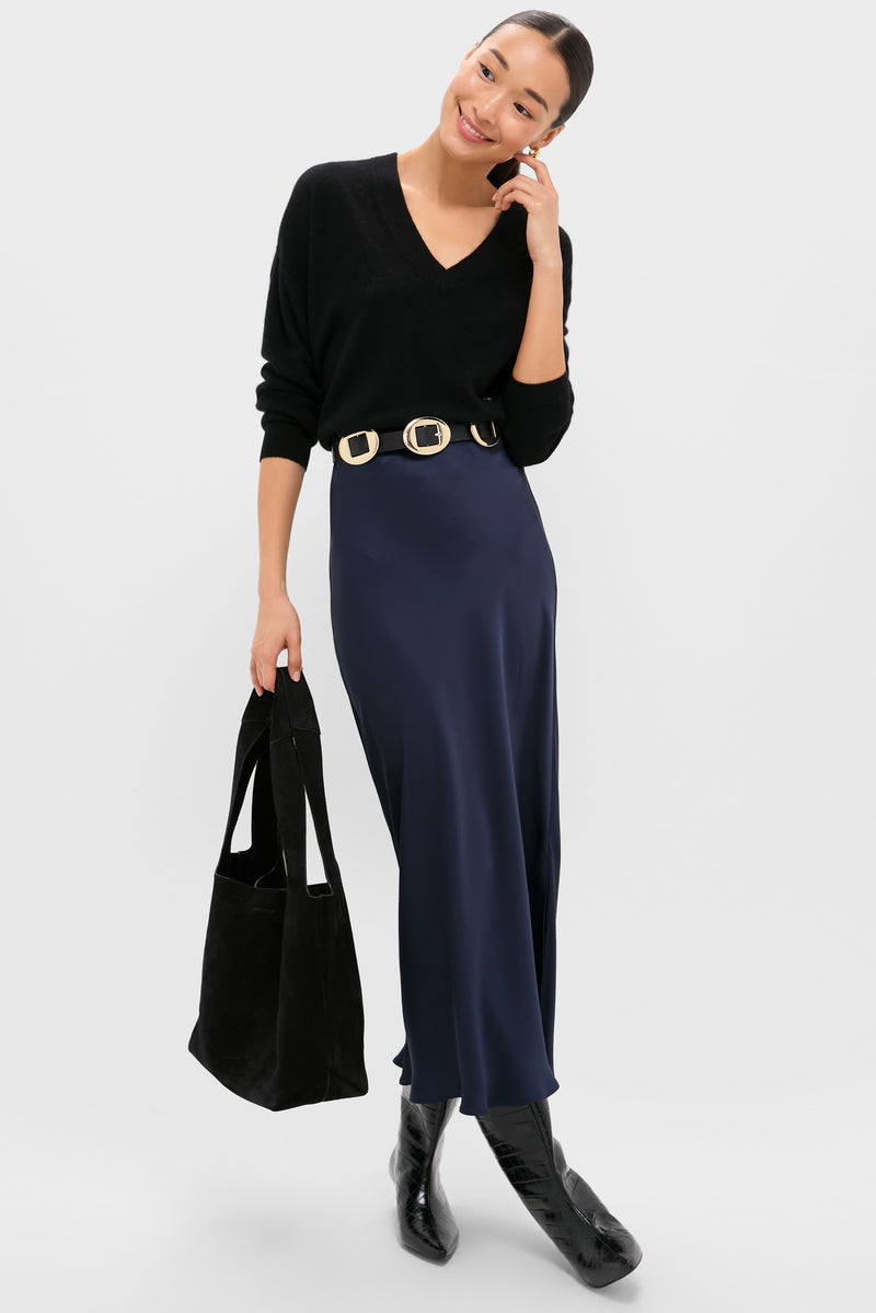 A woman smiles in a Tuckernuck oversized black cashmere V-neck sweater, navy blue satin skirt with wide belt, black boots, and carries a black tote bag.