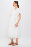 A woman in a white HATCH Carolina Dress with a scalloped, embroidered hem stands in profile, accessorized with gold jewelry and brown sandals—a chic maternity look against a plain white background.