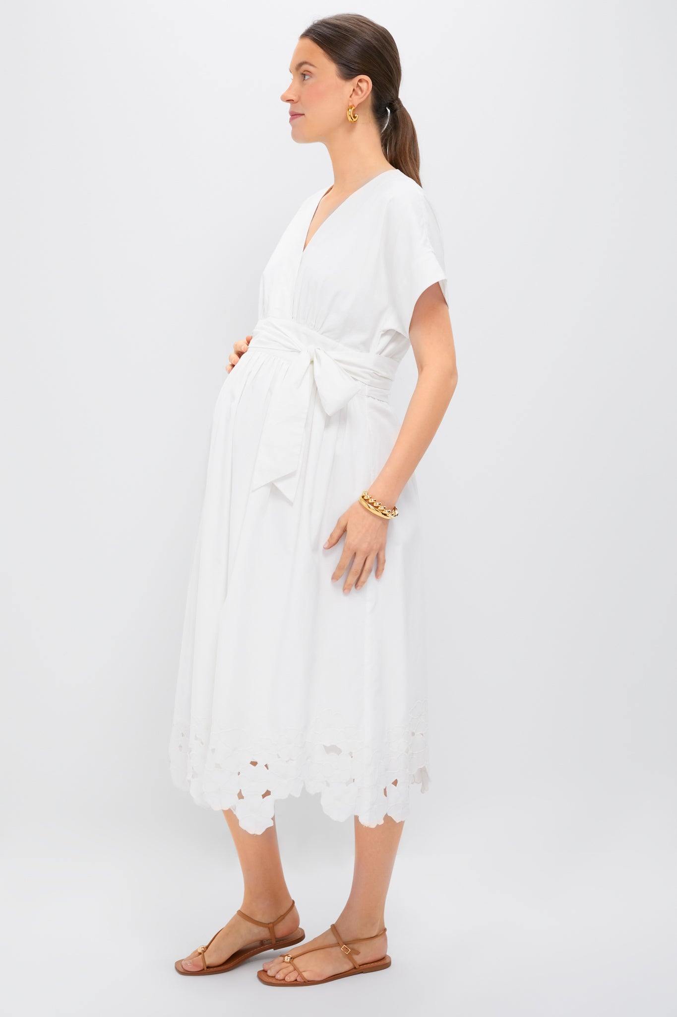 A woman in a white HATCH Carolina Dress with a scalloped, embroidered hem stands in profile, accessorized with gold jewelry and brown sandals—a chic maternity look against a plain white background.