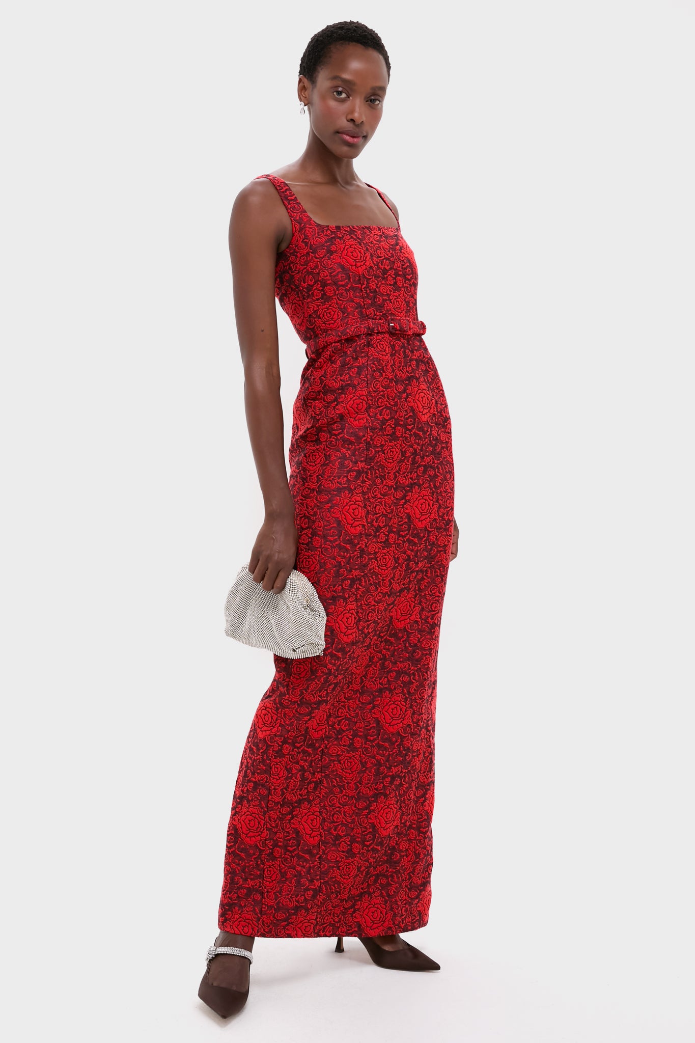 Model wearing a classic, high-end slim red dress with the effortless Chocolate Satin Mae Heels, featuring chic satin and jeweled ankle straps, holding a silver clutch.