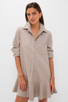 A woman models the Tuckernuck Callahan Shirt Dress in light beige, featuring long sleeves, a chest pocket, a ruffled hem, and a relaxed fit against a plain white background.