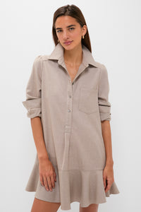 A woman models the Tuckernuck Callahan Shirt Dress in light beige, featuring long sleeves, a chest pocket, a ruffled hem, and a relaxed fit against a plain white background.