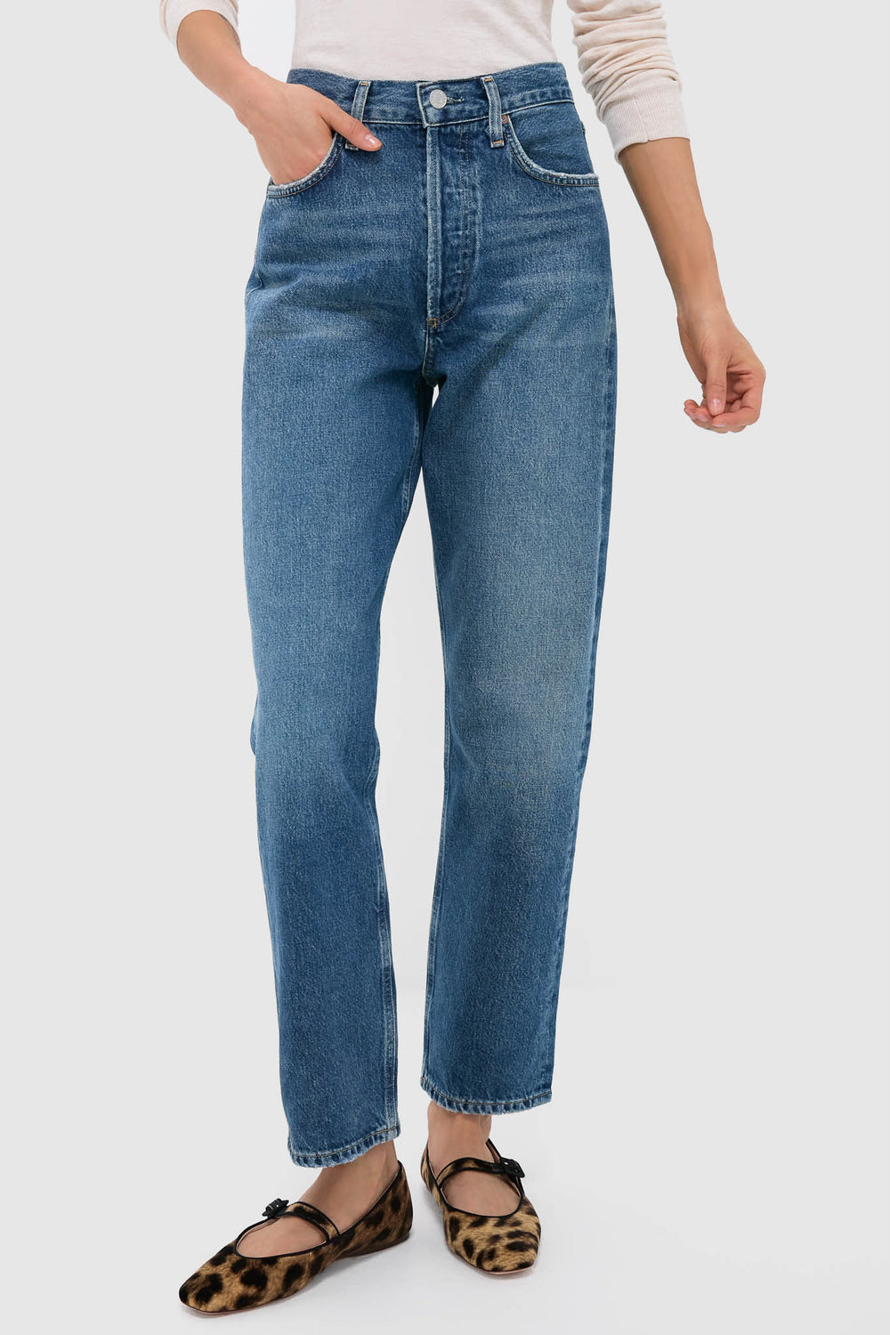 Someone stands against a plain light background wearing AGOLDE 90's straight leg blue jeans, a light long-sleeve top, and leopard print flat shoes.