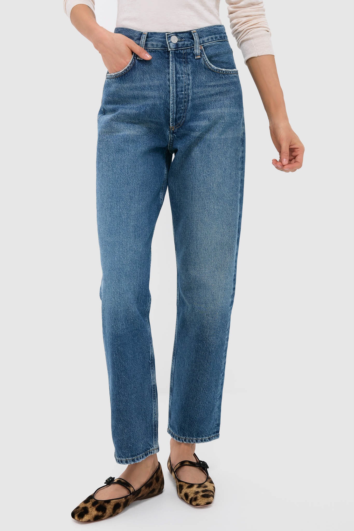 Someone stands against a plain light background wearing AGOLDE 90's straight leg blue jeans, a light long-sleeve top, and leopard print flat shoes.