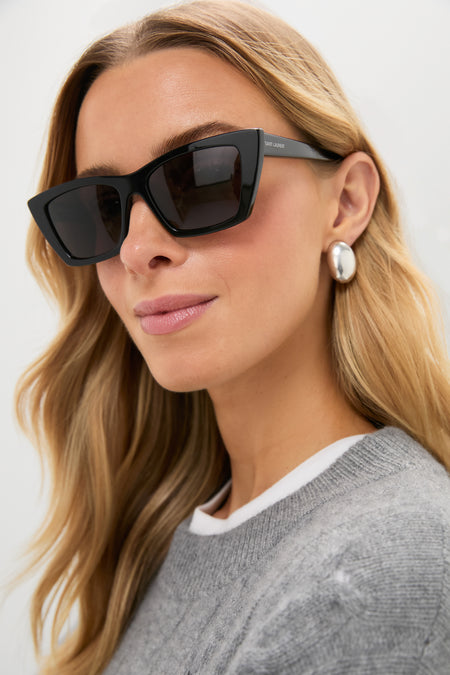 A woman with long blonde hair wears Saint Laurent cat-eye sunglasses, silver hoop earrings, and a grey sweater over a white top.