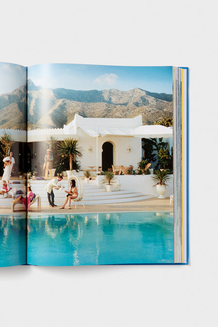 People unwind and mingle by a pool in front of a white villa with mountain views, as featured in a Harper Group photography book.
