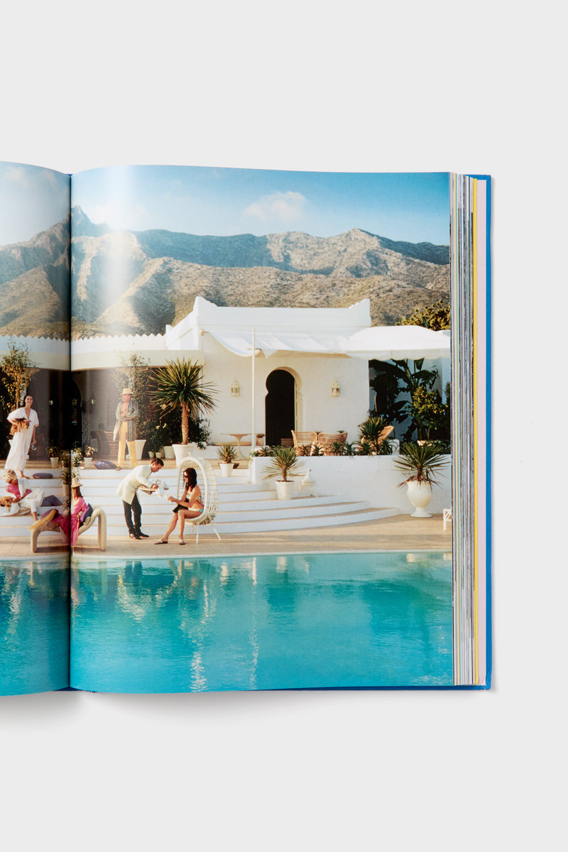 People unwind and mingle by a pool in front of a white villa with mountain views, as featured in a Harper Group photography book.