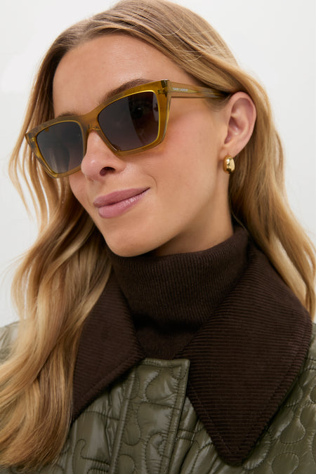Young woman wearing Shiny Transparent Ocra Mica Sunglasses with olive-green cat-eye frames, exuding a classic, effortless, and high-end style, paired with a fun, chic outfit.