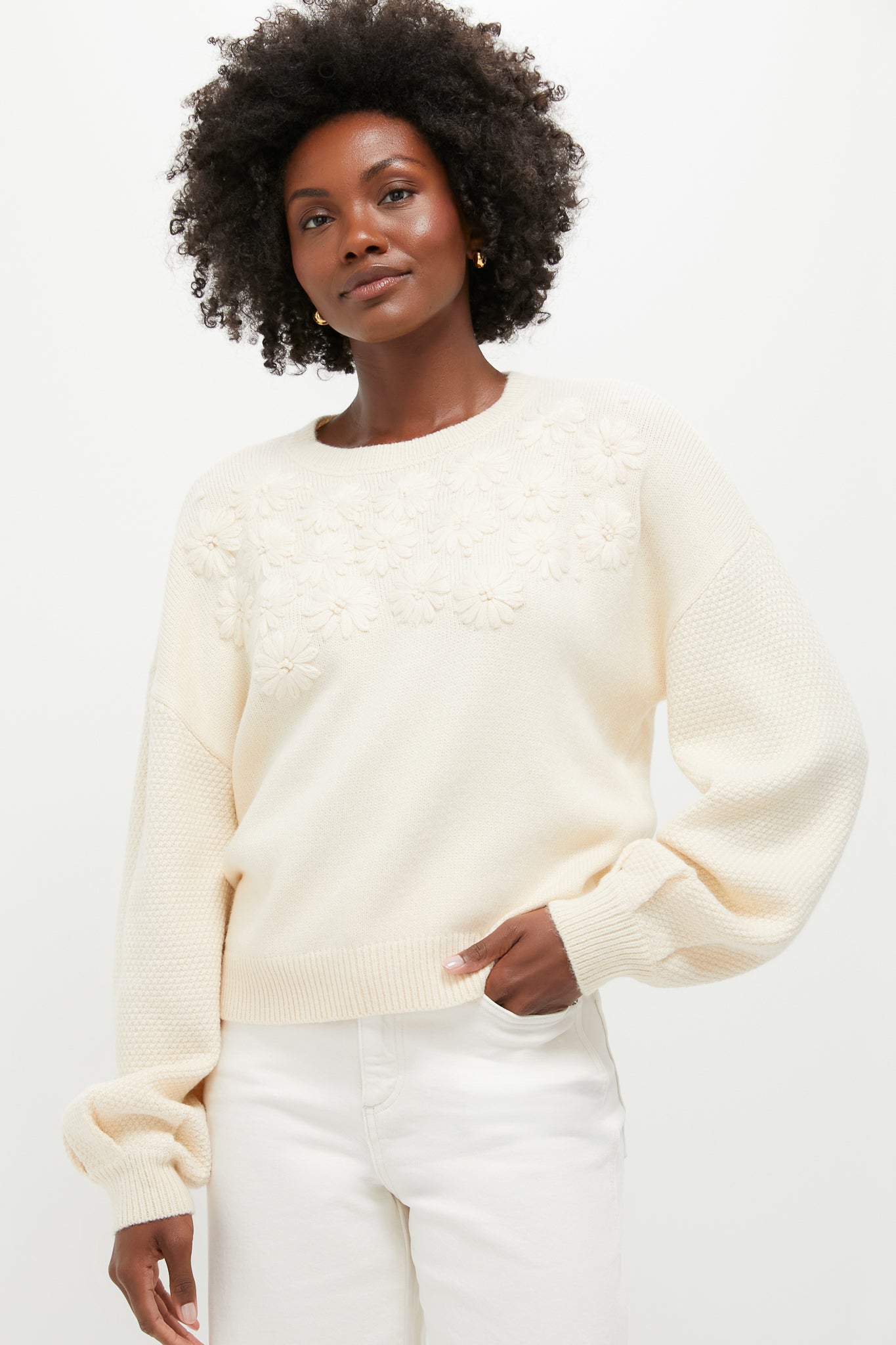 A woman stands confidently in a cream knit pullover with embroidered florals and bishop sleeves from Hyacinth House, paired with white pants.