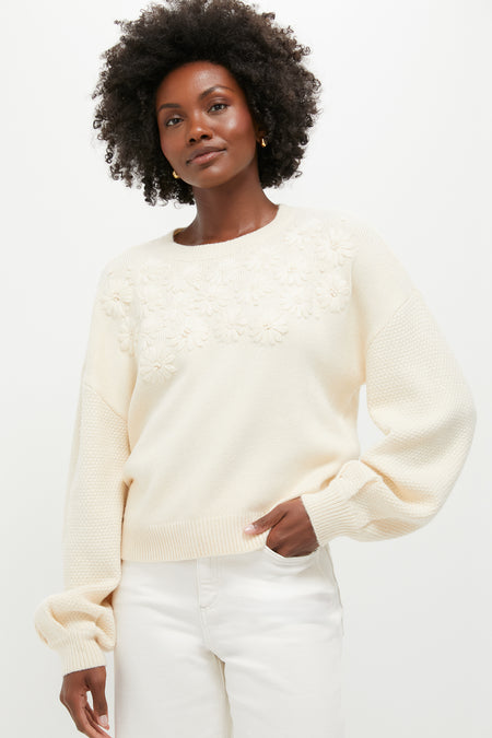 A woman stands confidently in a cream knit pullover with embroidered florals and bishop sleeves from Hyacinth House, paired with white pants.