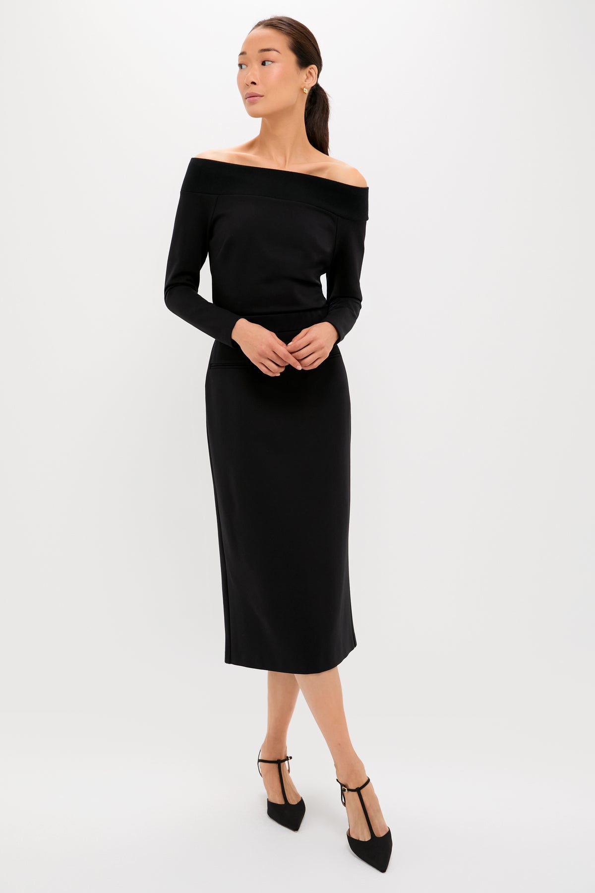 A woman poses against a plain background wearing a Tuckernuck black off-the-shoulder knit top and long-sleeve midi dress, creating a slim silhouette, paired with black pointed-toe heels featuring ankle straps.