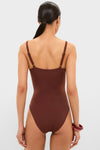 Back view of a woman in a classic, chocolate-brown Cappuccino Isabel One Piece swimsuit with adjustable straps, showcasing effortless style complemented by gold hoops and red bangles.