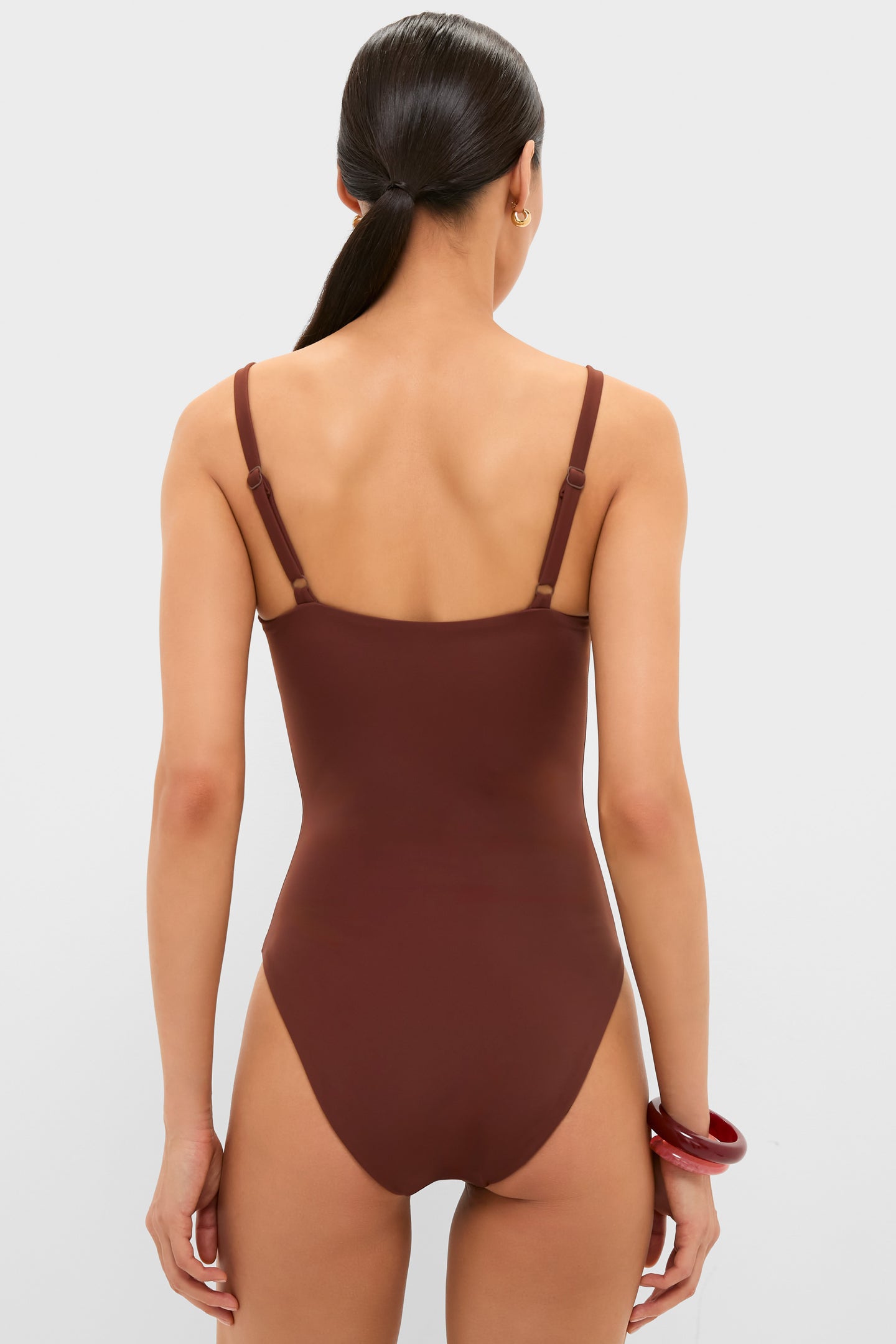 Back view of a woman in a classic, chocolate-brown Cappuccino Isabel One Piece swimsuit with adjustable straps, showcasing effortless style complemented by gold hoops and red bangles.