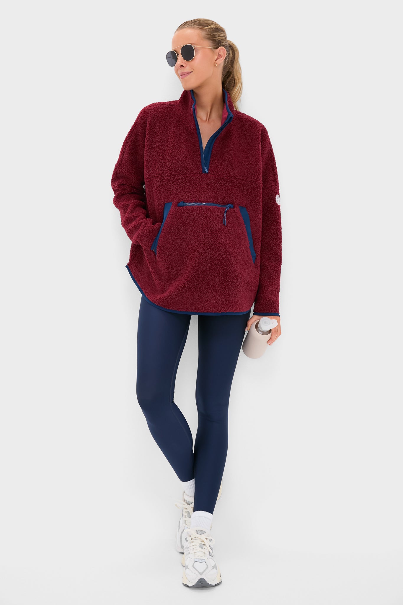 Woman wearing Cabernet Fleece Cady Quarter Zip pullover with navy trim, paired with leggings and sneakers, holding a water bottle, embodying classic, effortless, and fun style.