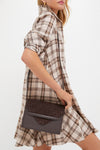 A woman in a beige and brown plaid dress holds a dark brown Tuckernuck Accessories Stevie crossbody at her side, highlighting a chic and hands-free bag option.
