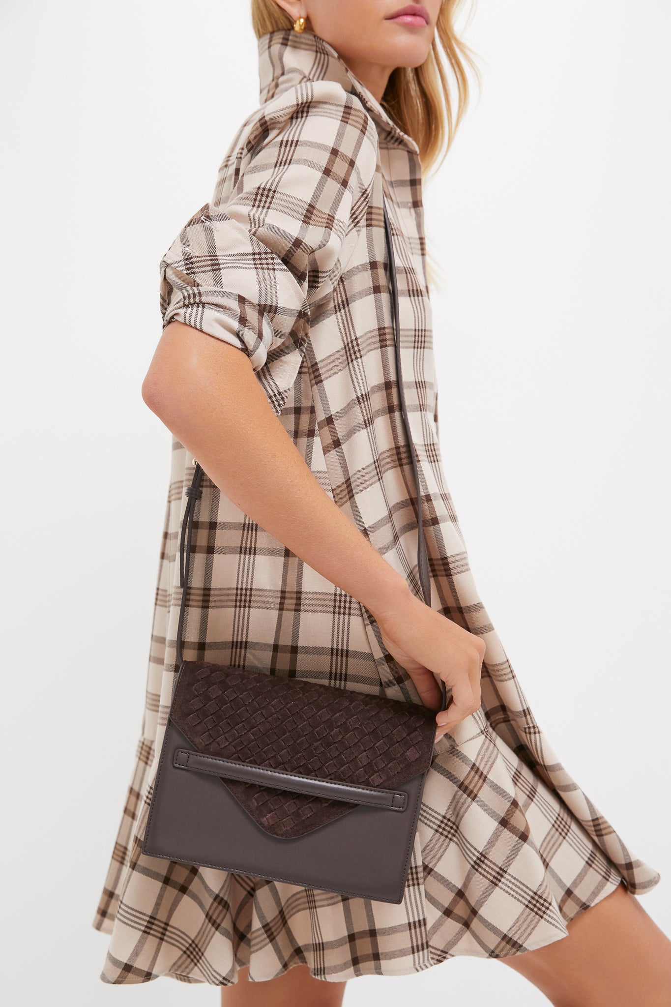 A woman in a beige and brown plaid dress holds a dark brown Tuckernuck Accessories Stevie crossbody at her side, highlighting a chic and hands-free bag option.