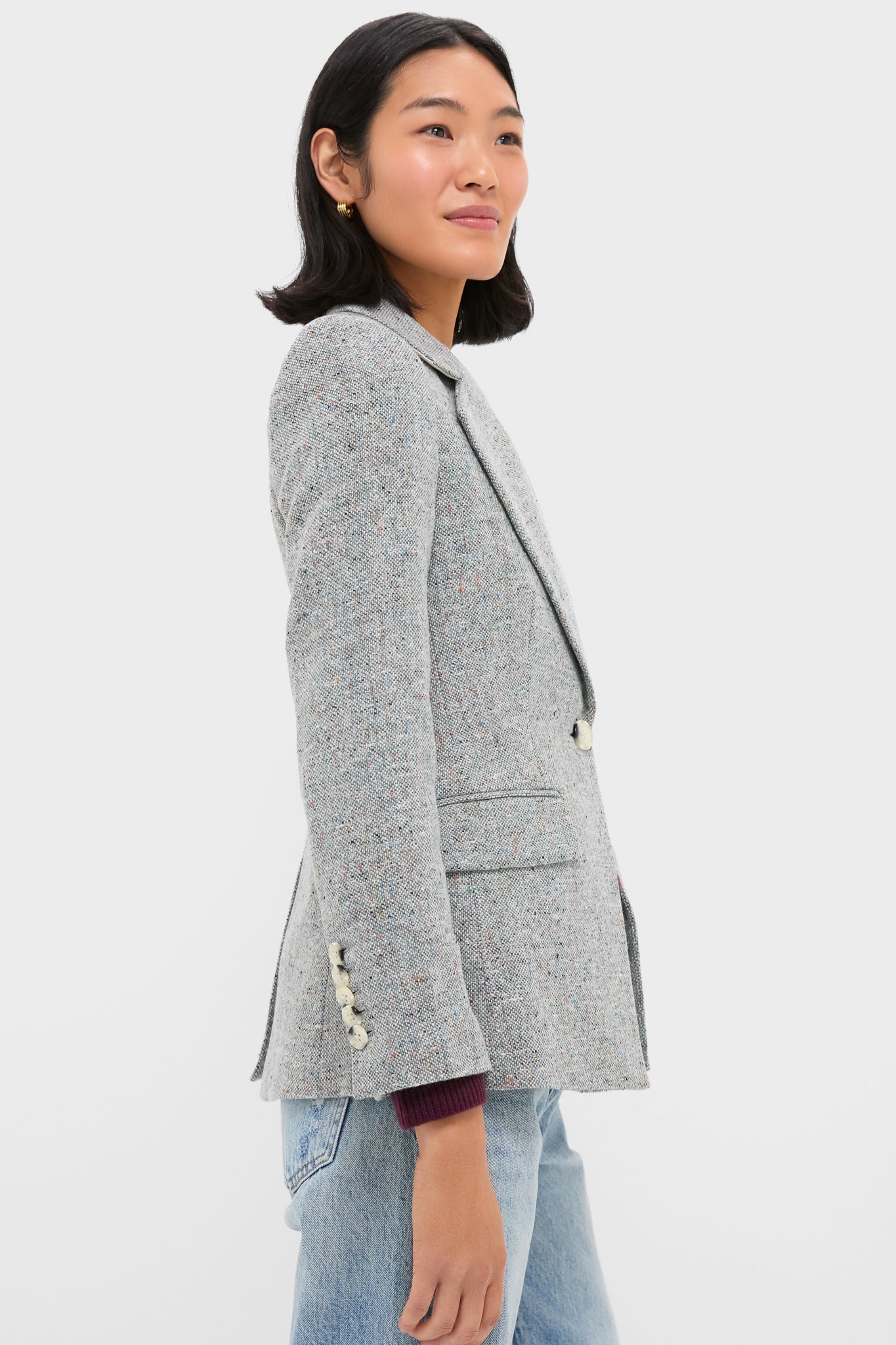 A woman stands in profile against a white background wearing a Veronica Beard light gray single-breasted blazer with pearl buttons and light blue jeans.