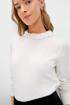 A woman stands against a plain light background, wearing the Tuckernuck Lina Sweater—a white long-sleeve pullover featuring a ruffled neckline.