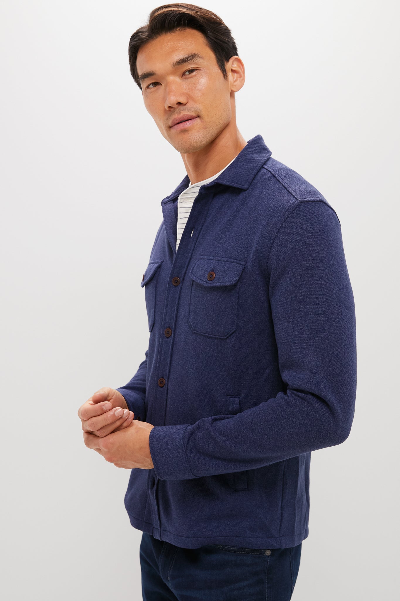 A man stands against a plain background, wearing a Peter Millar Navy Crown Sweater Shirt Jacket made from recycled performance fabric over a white striped shirt and dark jeans, looking slightly to the side.