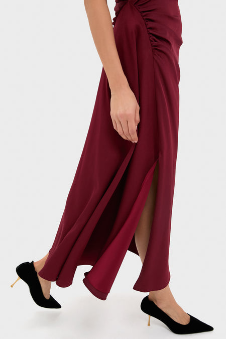 A person wearing a SIMKHAI oxblood midi dress with a side slit and black high heels walks, shown from the waist down—elegant occasion wear in motion.