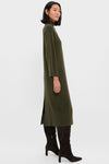 A woman stands in profile wearing the Pomander Place Vivianne Dress, an olive green turtleneck style with a high neck, paired with knee-high dark brown suede boots.