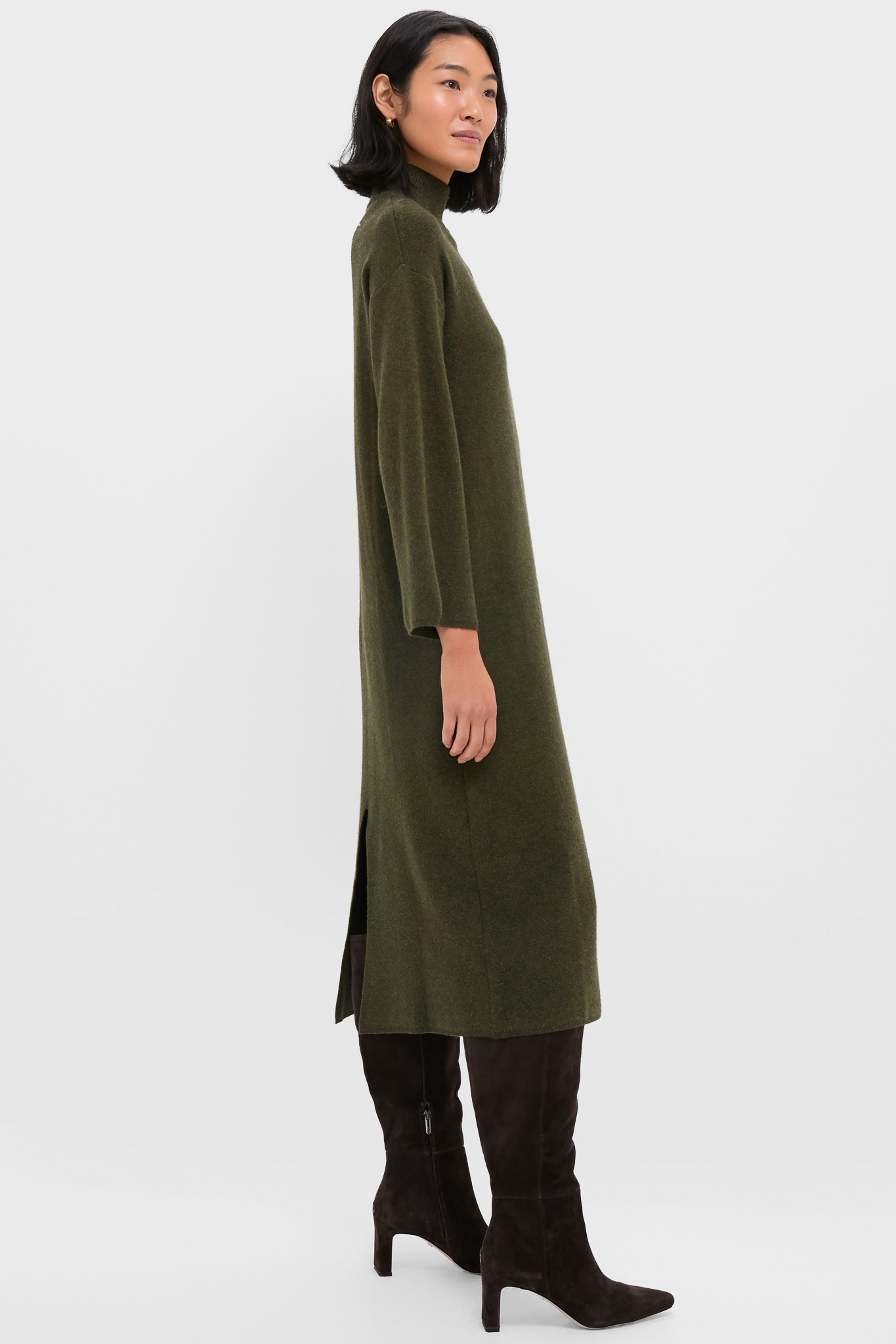A woman stands in profile wearing the Pomander Place Vivianne Dress, an olive green turtleneck style with a high neck, paired with knee-high dark brown suede boots.