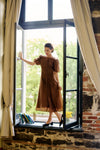 Woman standing in a windowsill in a brown midi dress and black mules. 