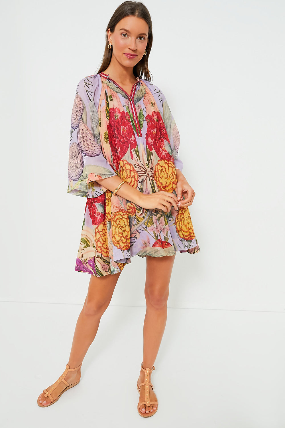 A woman wears the Carolina K Floral Lilac Amala Dress with tan sandals, featuring a loose-fitting cotton mini and vibrant floral prints.