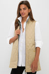 A woman stands against a white background wearing a Barbour x Tuckernuck beige quilted vest with a reversible paisley liner over a white button-up shirt and black pants.