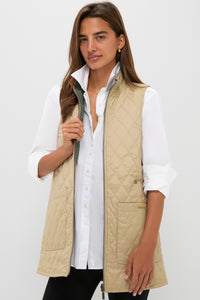 A woman stands against a white background wearing a Barbour x Tuckernuck beige quilted vest with a reversible paisley liner over a white button-up shirt and black pants.
