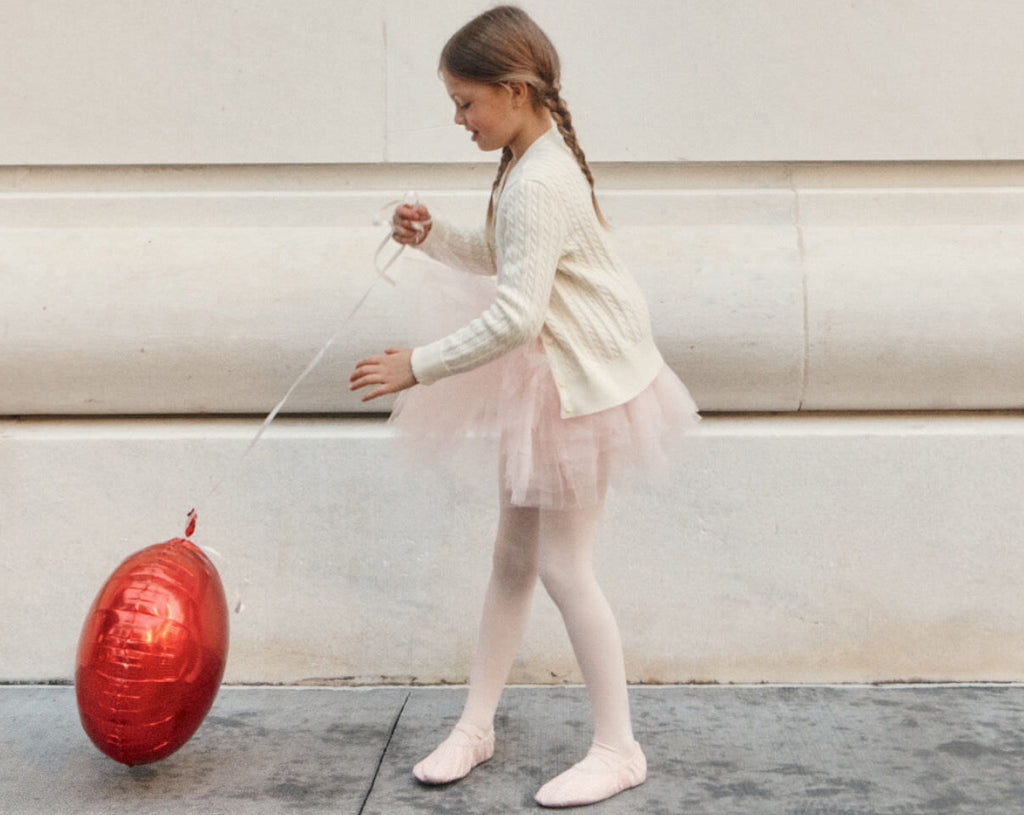 A little girl wears pink ballet tights, pink ballet shoes, a pink tutu, and a white cable knit cardigan while playing with a red balloon.