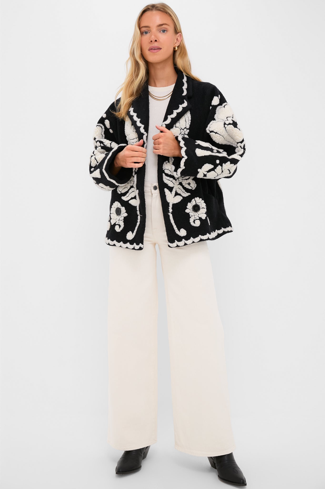 Blonde woman in a classic Black Cream Floral Arabesque Fleece Coat with scalloped trim, styled effortlessly over cream trousers and black ankle boots for a high-end, fun look.