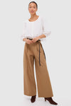 Woman models classic Umber Slubby Cotton Atelier Pleated Pant 2, styled effortlessly with a crisp blouse, black belt, and heeled boots, embodying high-end, fun sophistication.