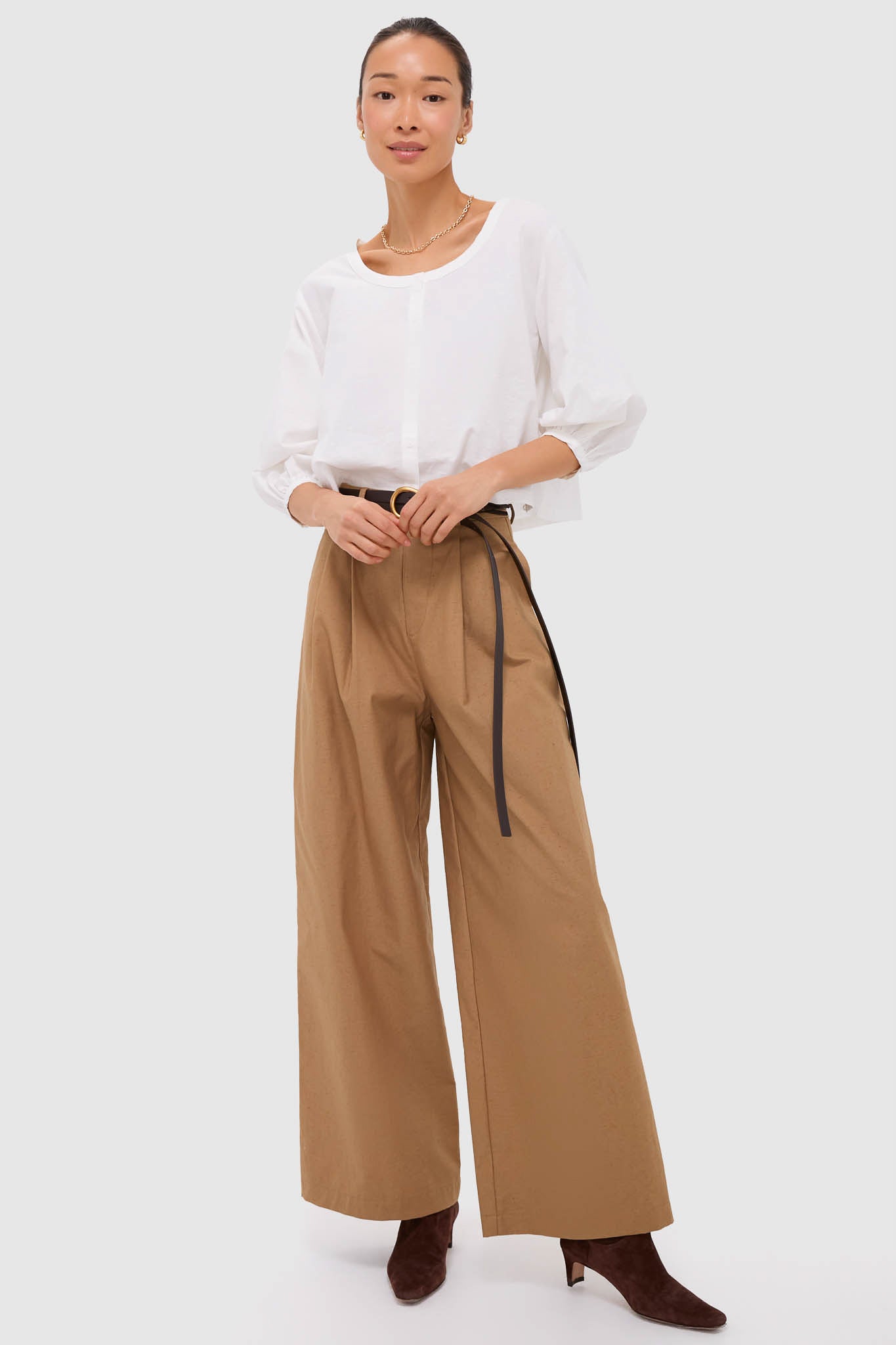 Woman models classic Umber Slubby Cotton Atelier Pleated Pant 2, styled effortlessly with a crisp blouse, black belt, and heeled boots, embodying high-end, fun sophistication.