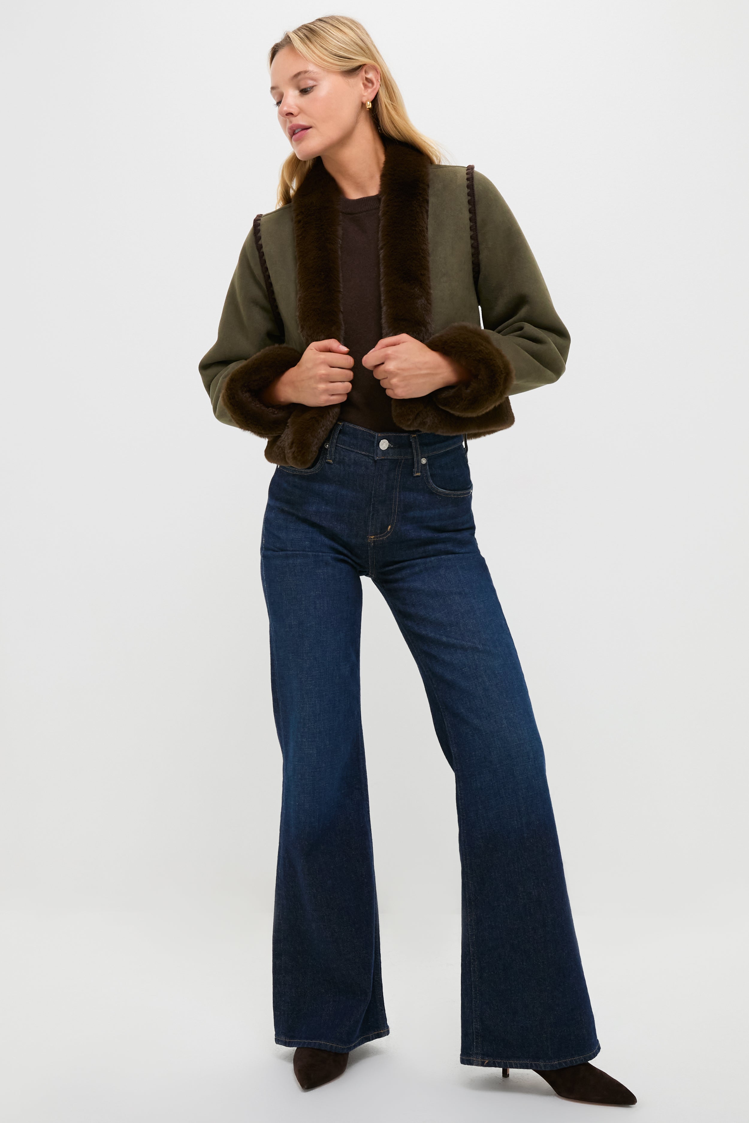 A woman wears a Hyacinth House olive faux suede cropped jacket with dark faux fur trim, a brown top, wide-leg blue jeans, and heeled boots, standing against a plain white background.