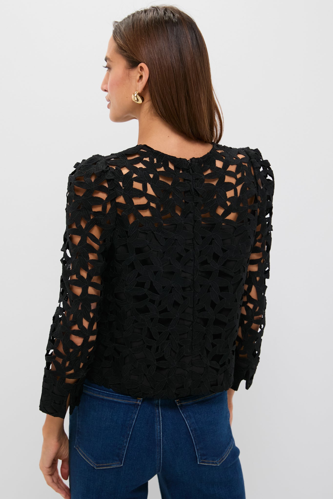 Back view of a woman in a classic Black Lace Elodie Blouse with floral lace cutouts, effortlessly paired with jeans and gold hoop earrings for a high-end, fun look.