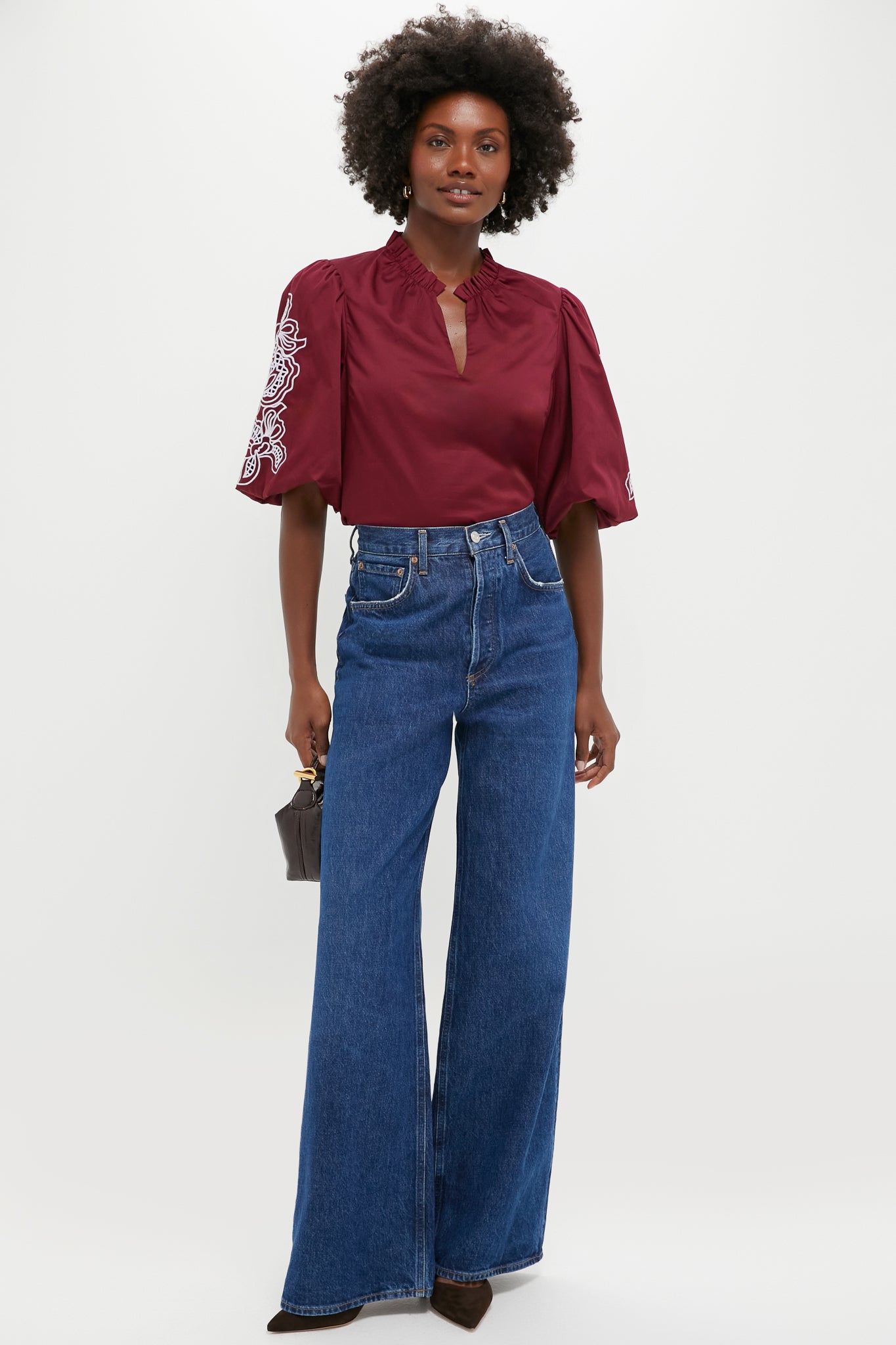 A woman wears a Hyacinth House burgundy embroidered blouse with puff sleeves, high-waisted wide-leg blue jeans, brown heels, and holds a small handbag against a plain background.