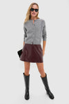 Model wearing the classic Oxblood Meeghan Mini Skirt, an effortless high-end vegan leather piece, paired with a gray cardigan, knee-high boots, and a black clutch.