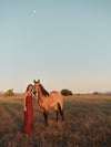 Woman wearing a sleeveless maxi dress with eyelet details in a rust color standing next to a horse. 