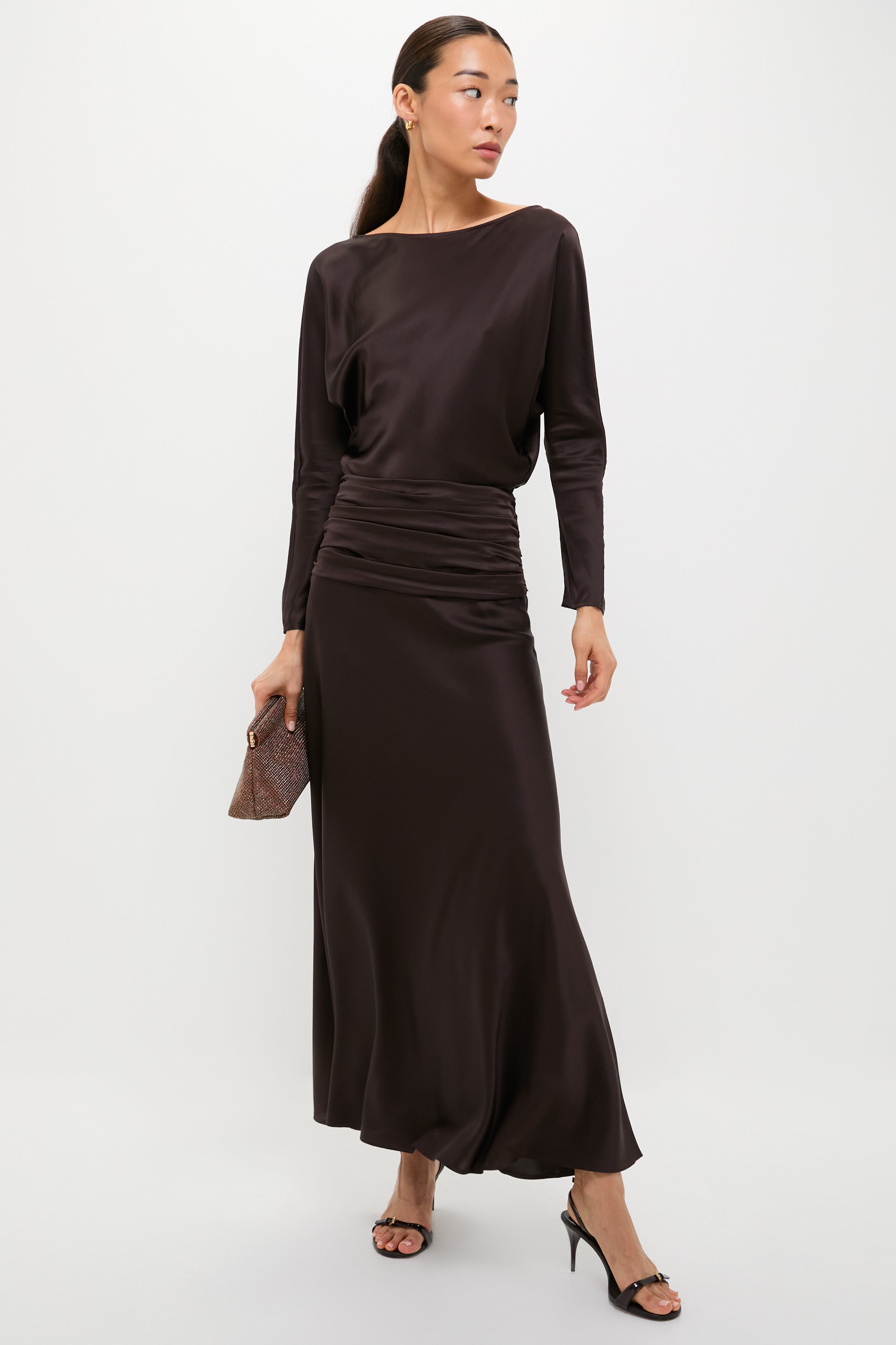 A woman models a STAUD long sleeve dress with a ruched waist, perfect for occasion dressing, paired with a small clutch and black heeled sandals, standing against a plain white background.