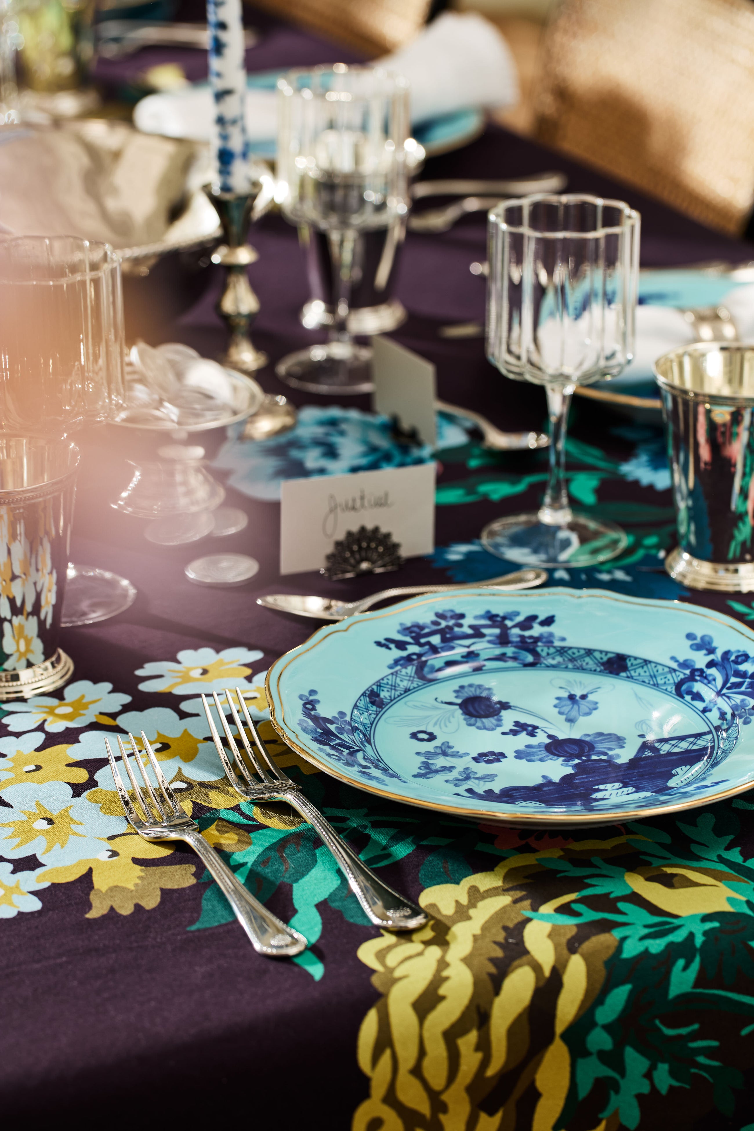A close-up of a table setting featuring Ginori 1735 handcrafted blue floral porcelain plates, silverware, clear and silver glasses, and a yellow, green, and black floral tablecloth.