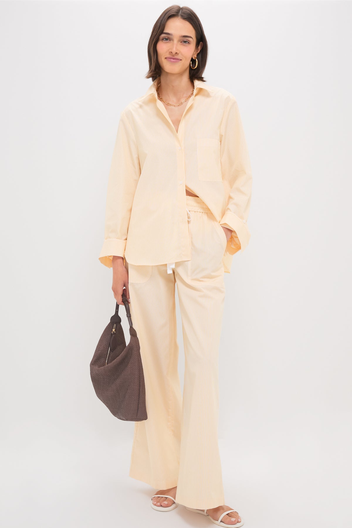 A person stands against a plain background wearing the TWP Next Morning Shirt in Ivory and Yellow with an oversized fit, paired with matching wide-leg pants. The look is completed with a dark brown bag, white sandals, and is made from 100% cotton.