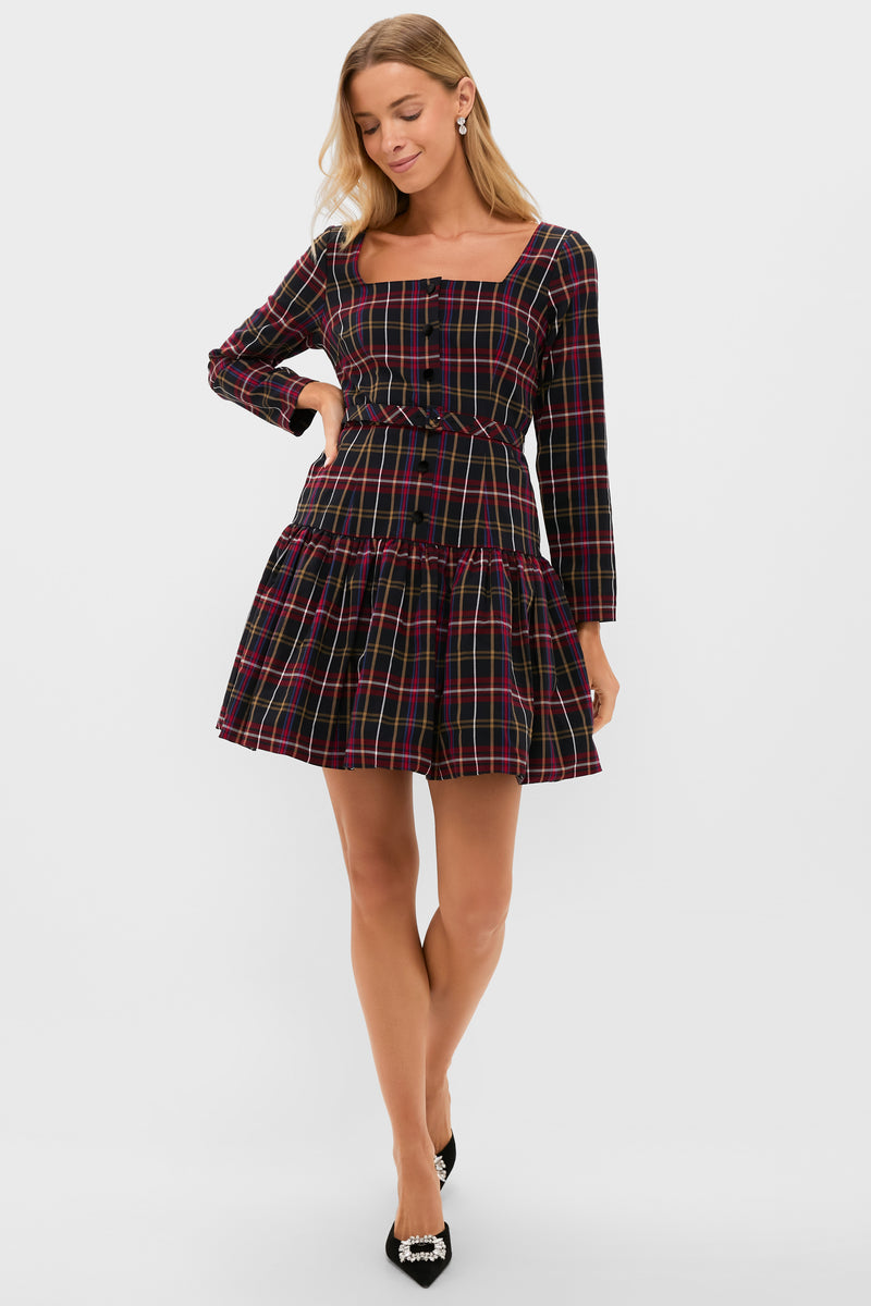 A woman wearing the Tuckernuck black tartan mini dress with long sleeves, a square neckline, and a tiered skirt stands with one hand on her hip, looking down—a chic choice for festive occasions.