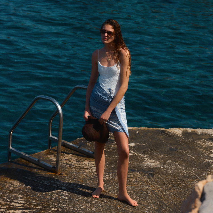 Woman standing by the water in a white one piece and a blue pareo wrapped around her waist