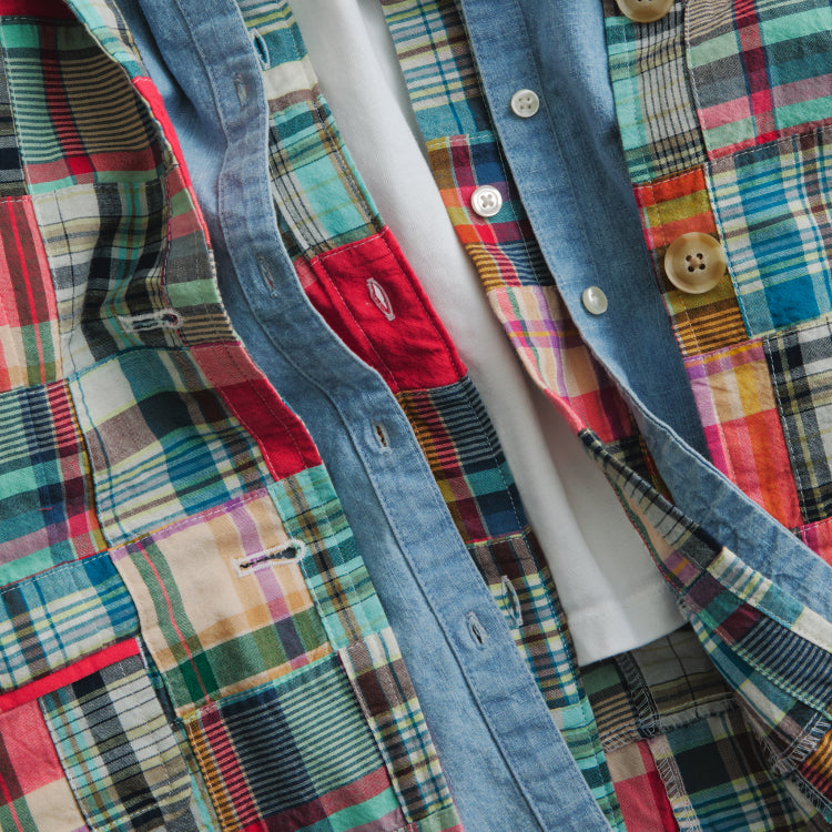 Several shirts layered together. The base is a white tee and then three shirts are layered on top of it. A madras shirt with white buttons, a chambray shirt with white buttons, and a madras shirt with chambray buttons. 