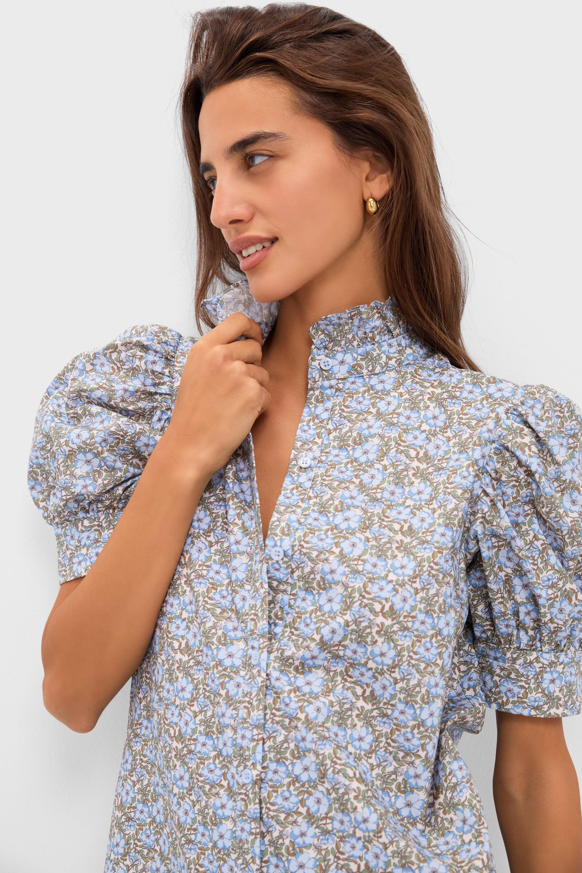 A woman models a Hyacinth House light blue floral blouse with puffed sleeves and a ruffle collar, gazing to the side while holding the collar against a plain background.
