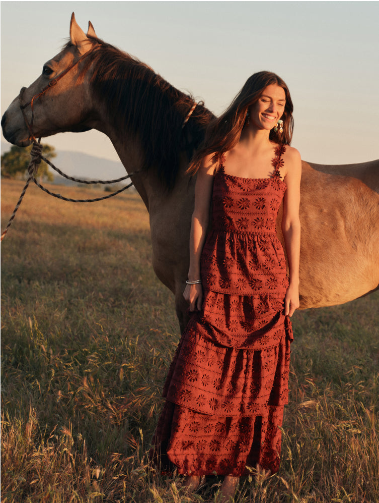 Woman in a rust colored maxi dress with eyelet details standing in front of a horse. 